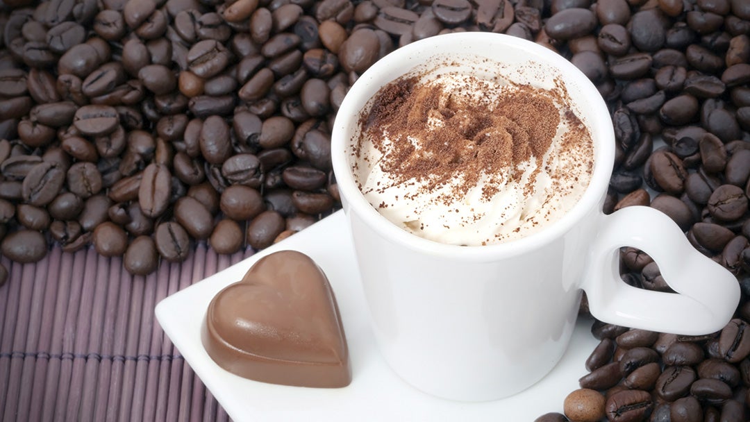 Taza de café moka encima de granos de café con un bombón con forma de corazón al costado