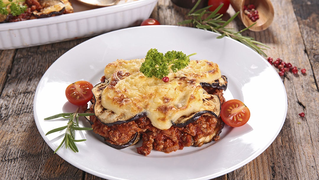 Lasaña de berenjenas con queso gratinado, tomates cherry y hierbas, en un plato sobre madera.