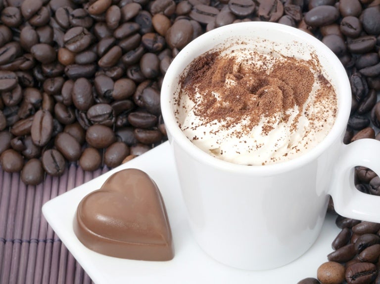 Taza de café moka encima de granos de café con un bombón con forma de corazón al costado