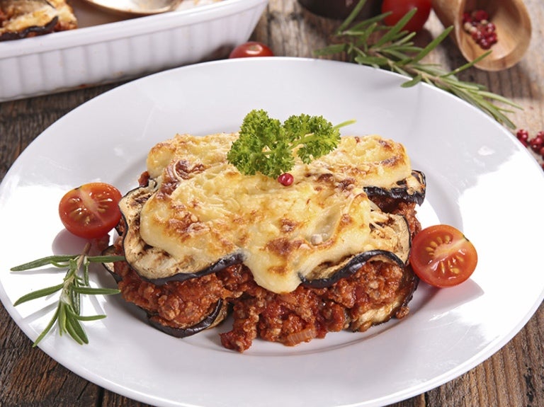 Lasaña de berenjenas con queso gratinado, tomates cherry y hierbas, en un plato sobre madera.